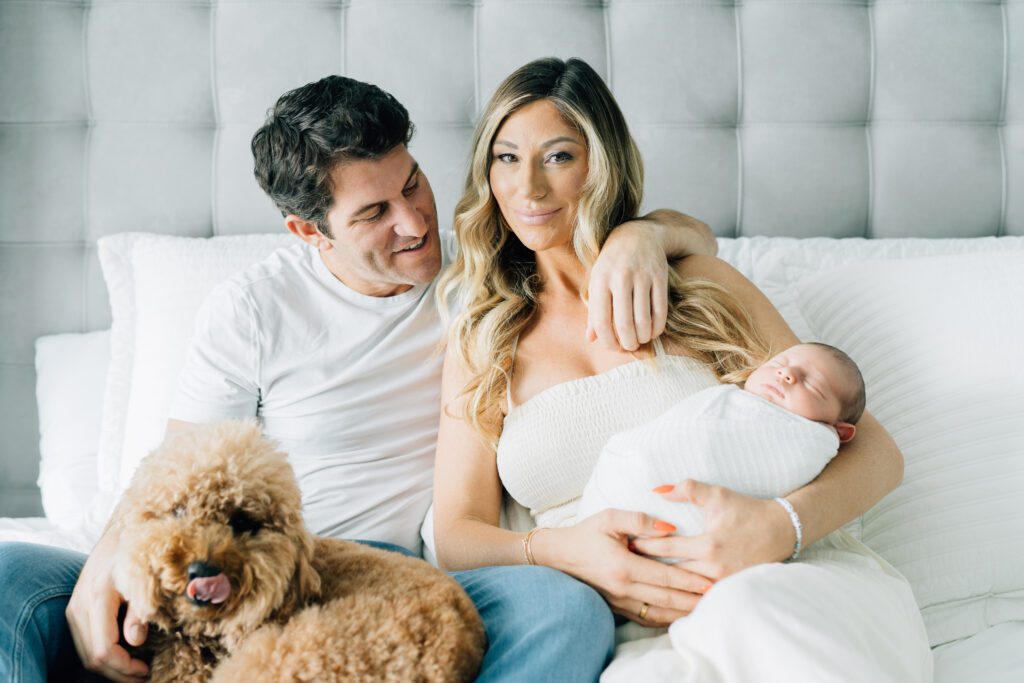 mom and dad sitting on a bed with newborn baby and dog