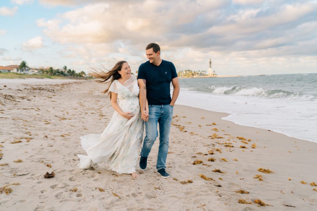  couple walking down the beach in deerfield during sunset time. mom is pregnant and dad is admiring her. hillsboro inlet lighthouse in the background.