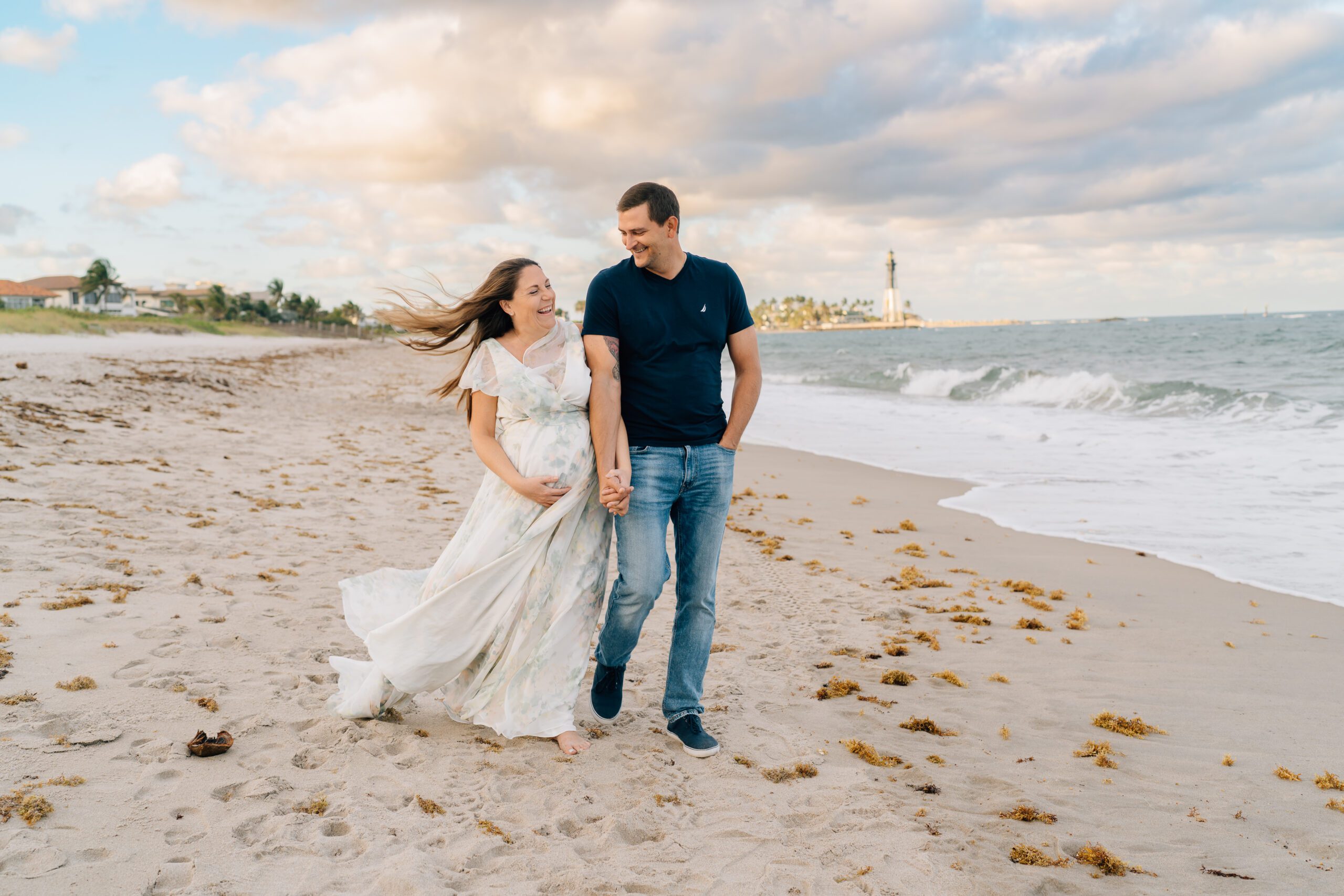 couple walking down the beach in deerfield during sunset time. mom is pregnant and dad is admiring her. hillsboro inlet lighthouse in the background.